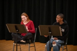 Two actors seated at music stands laugh together during rehearsal