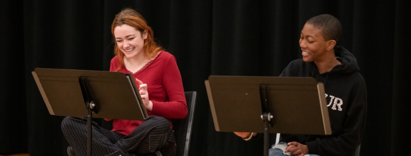 Two actors seated at music stands laugh together during rehearsal