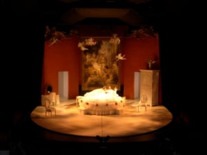 Two actors lay in an ornate bed surrounded by classical furniture in The Misanthrope