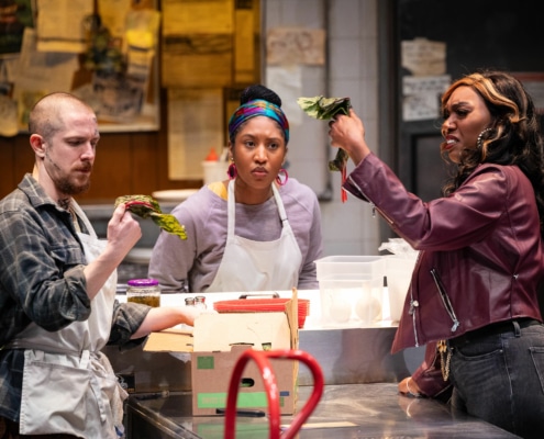 Two cooks wearing aprons and a woman in a purple leather jacket examine a wilted leafy green vegetable with confused expressions on their faces.