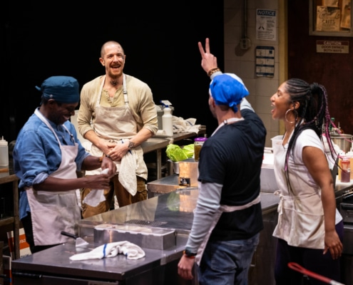 Four cooks wearing aprons celebrate in the kitchen. One man with his back to the camera holds up a peace sign.