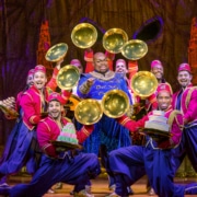 Genie points to the audience surrounded by a group of colorfully dressed dancers. The dancers show off desserts from beneath golden cloches