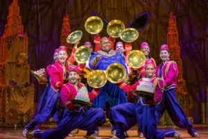 Genie points to the audience surrounded by a group of colorfully dressed dancers. The dancers show off desserts from beneath golden cloches