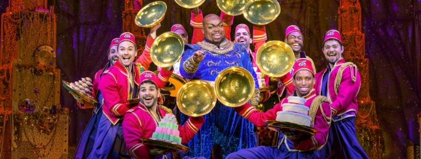 Genie points to the audience surrounded by a group of colorfully dressed dancers. The dancers show off desserts from beneath golden cloches