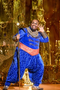 Genie, wearing a bright blue costume and holding an old fashioned microphone stand, gestures to his ear as if he can't hear you