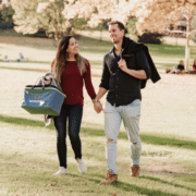 A couple holds hands while walking through a park with a insulated bag for a picnic