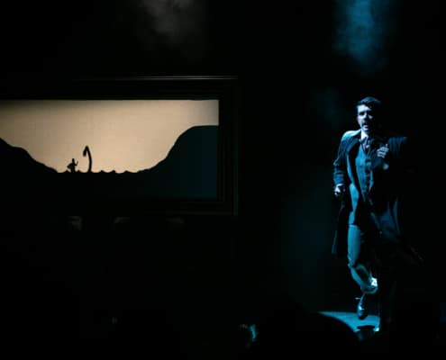 Richard Hannay runs on a dark stage, shadow puppets on the screen behind him