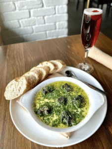 A plate of buttery escargot with a side of sliced baguette. A sparkling, red cocktail in a champagne flute accompanies.