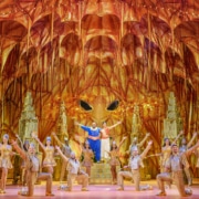 Genie and Aladdin stand with their arms around each other center stage. The company of Aladdin poses with an arm extended around the glittering, gold stage, presenting the scene to the audience