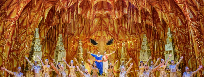 Genie and Aladdin stand with their arms around each other center stage. The company of Aladdin poses with an arm extended around the glittering, gold stage, presenting the scene to the audience