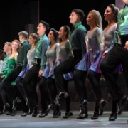 A row of smiling dancers wearing green and black costumes performing in Riverdance