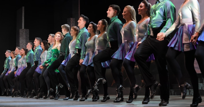A row of smiling dancers wearing green and black costumes performing in Riverdance
