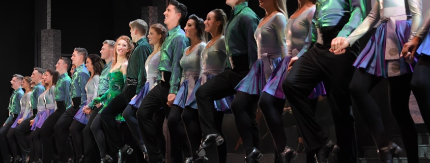 A row of smiling dancers wearing green and black costumes performing in Riverdance