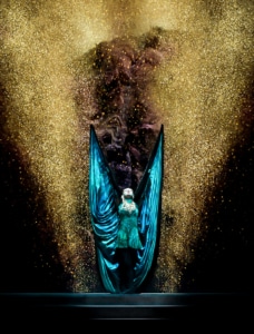 A woman wearing long silks, like wings, holds them up in a shower of gold glitter in Riverdance