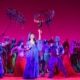 Jasmine dressed in a cape with a hood, walks through town, admiring a hanging lantern in Aladdin.