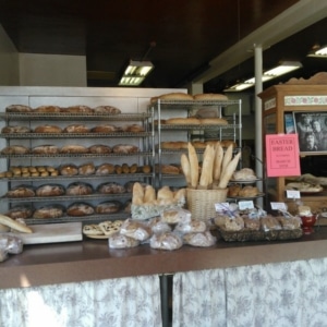 The checkout counter at Denver Bread Company is covered in loaves of bread for sale. Behind, there are cooling racks filled with more loaves.