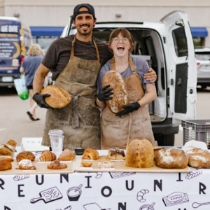 A couple smiles, posing for a photo at their farmers market booth, holding loaves of bread