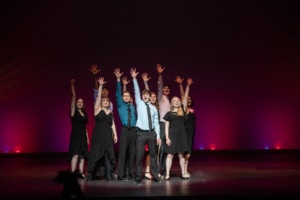 The Lead Performer nominees stand huddled together, singing the final note of the medley at the Bobby G Awards. Each has one arm raised, fingers reaching high.