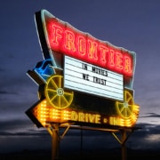 The Frontier Drive-Inn sign, lit up at dusk. It reads 'In Movies We Trust.'