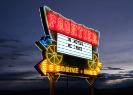 The Frontier Drive-Inn sign, lit up at dusk. It reads 'In Movies We Trust.'