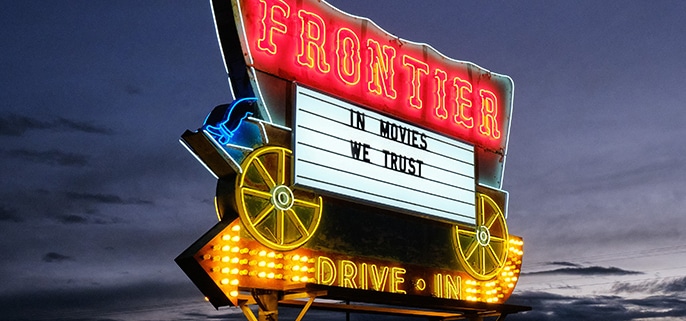 The Frontier Drive-Inn sign, lit up at dusk. It reads 'In Movies We Trust.'