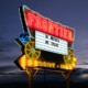 The Frontier Drive-Inn sign, lit up at dusk. It reads 'In Movies We Trust.'