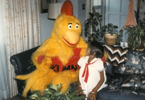 A young girl sits with a chicken mascot in her living room