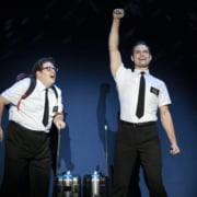 Two actors in the Book of Mormon stand excitedly onstage, wearing the classic white, short sleeved button down, black tie, and nametag