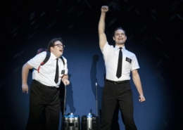 Two actors in the Book of Mormon stand excitedly onstage, wearing the classic white, short sleeved button down, black tie, and nametag