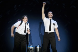 Two actors in the Book of Mormon stand excitedly onstage, wearing the classic white, short sleeved button down, black tie, and nametag