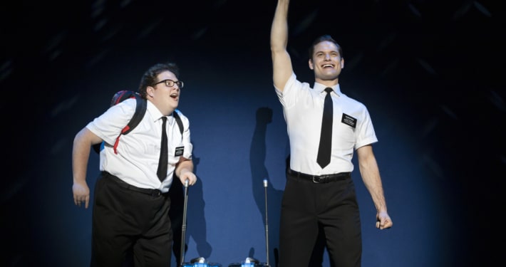 Two actors in the Book of Mormon stand excitedly onstage, wearing the classic white, short sleeved button down, black tie, and nametag