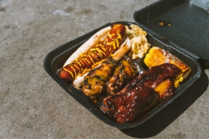 Wings, a loaded hotdog and more are overflowing from a black takeout container