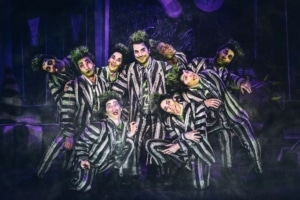 Beetlejuice, wearing his signature black and white striped suit, is surrounded by matching dancers imitating his insane expression