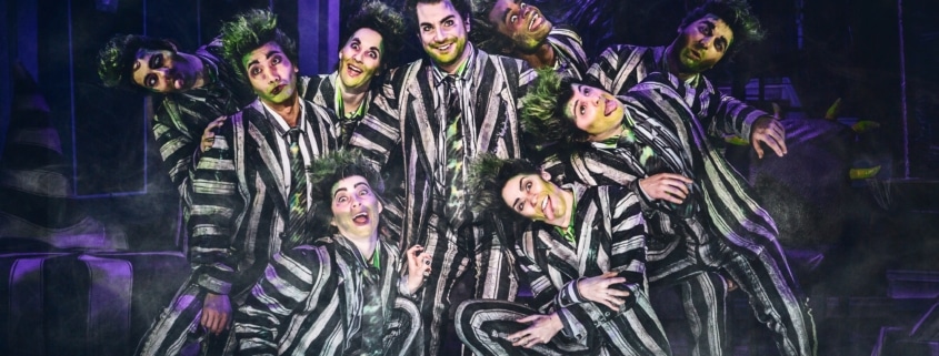 Beetlejuice, wearing his signature black and white striped suit, is surrounded by matching dancers imitating his insane expression