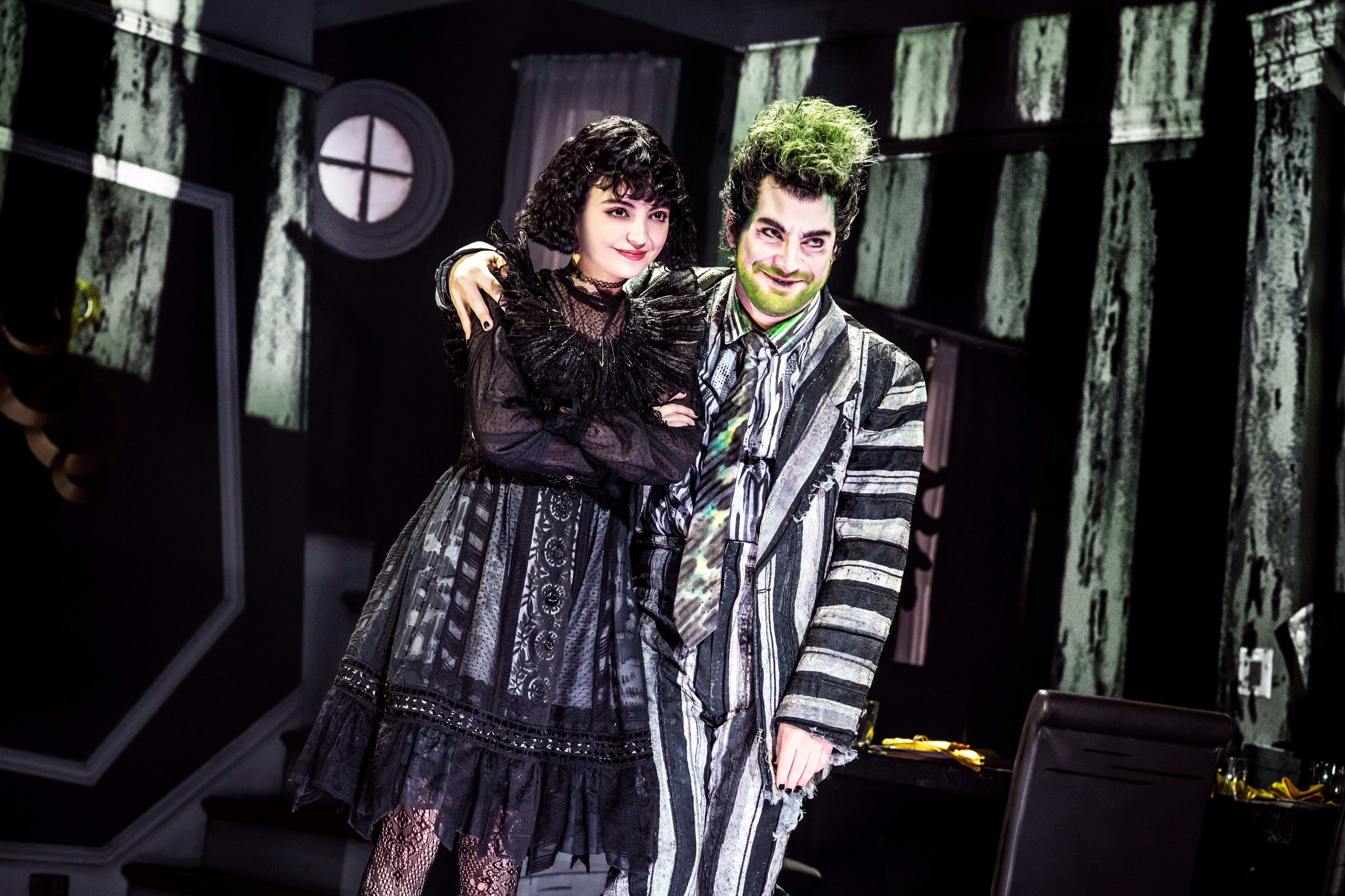 Lydia and Beetlejuice stand together, his arm around her shoulders. She is wearing a Gothic black dress, he is wearing his famous striped suit.