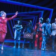 Juno stands apart, her arms raised, as the cast of Beetlejuice looks on. Juno is wearing a red printed skirt suit.