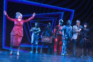 Juno stands apart, her arms raised, as the cast of Beetlejuice looks on. Juno is wearing a red printed skirt suit.