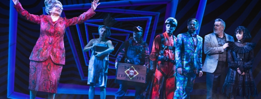 Juno stands apart, her arms raised, as the cast of Beetlejuice looks on. Juno is wearing a red printed skirt suit.