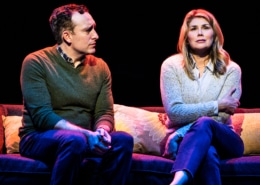 A man and woman sit together on a couch. The woman is sitting with her arms crossed, closed off. The man looks sad, watching her.