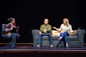 A man and woman sit on a couch, in therapy. The therapist looks concerned while the woman speaks, her arms extended.