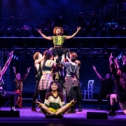 A group of dancers perform onstage. One dancer is lifted high with her arms outstretched. Others are kneeling, facing her.