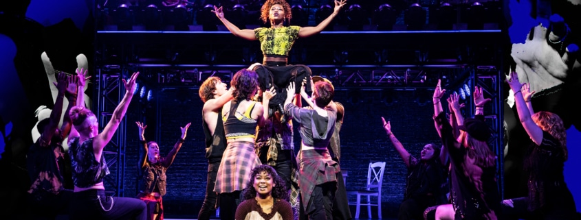 A group of dancers perform onstage. One dancer is lifted high with her arms outstretched. Others are kneeling, facing her.