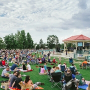 Hundreds of people sit on the grass, on blankets and in chairs. There is a band playing in a small gazebo.