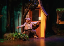 The Wolf poses in front of Grandma's house as he sings. He's bent on one knee with his arms outstretched.