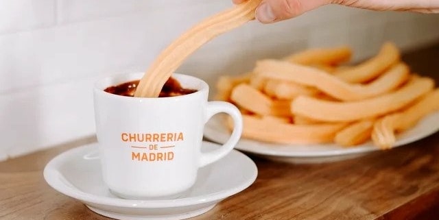 A plate of churros is in the background, and one is being dipped into a Churreria de Madrid mug filled with warm chocolate.