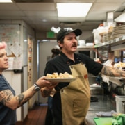 A man hands plates filled with food over to a woman. They are in the kitchen of Steuben's