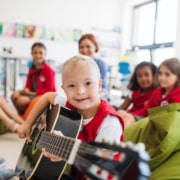 A down-syndrome boy with school kids and teacher sitting on the floor in class, playing guitar.