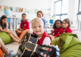 A down-syndrome boy with school kids and teacher sitting on the floor in class, playing guitar.