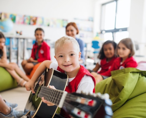 A down-syndrome boy with school kids and teacher sitting on the floor in class, playing guitar.