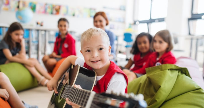A down-syndrome boy with school kids and teacher sitting on the floor in class, playing guitar.